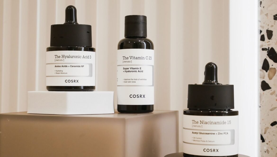 Three COSRX skincare serums displayed on modern geometric blocks: The Hyaluronic Acid 3, The Vitamin C 23, and The Niacinamide 15.