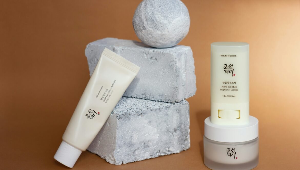 A display of Beauty of Joseon skincare products, including a sunscreen tube, matte sun stick, and moisturizer, arranged against rough gray concrete blocks and a warm brown background.