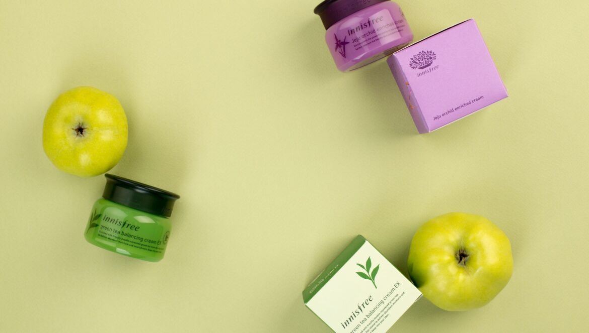 Innisfree skincare products, including green tea balancing cream and Jeju orchid enriched cream jars alongside their matching boxes and fresh green apples, arranged on a light green background.