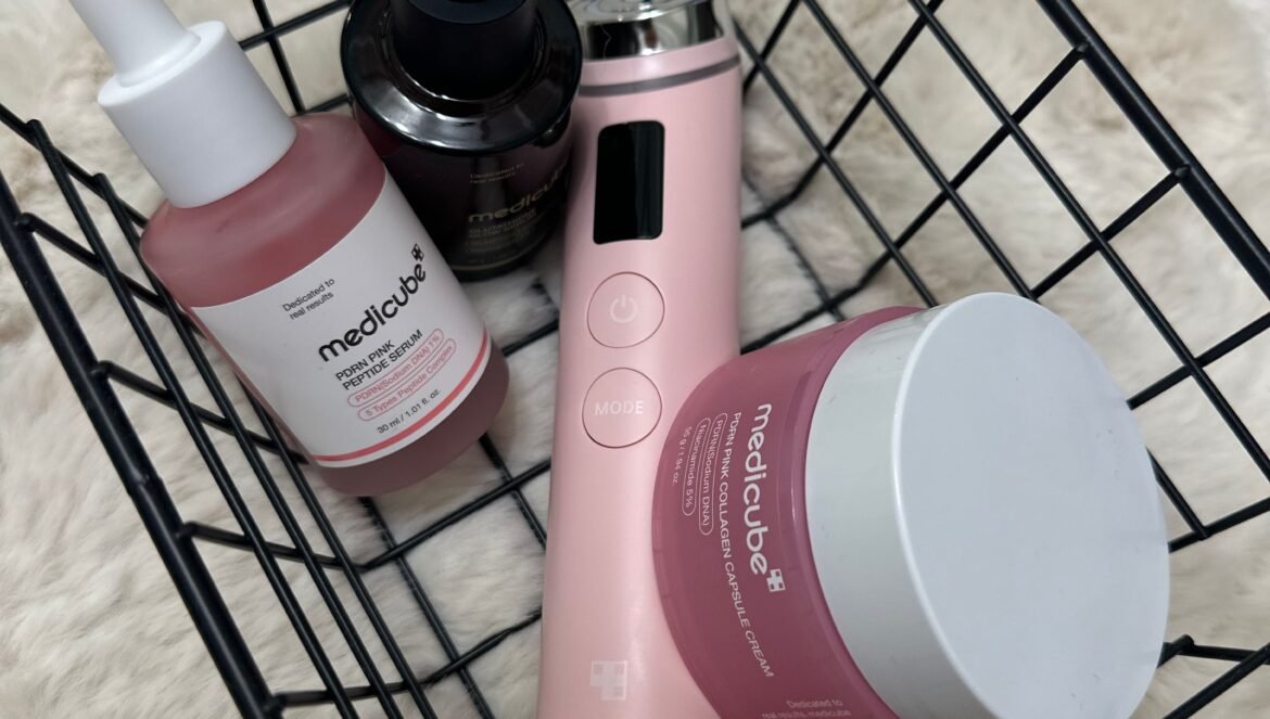 A black wire basket holding Medicube skincare products, including a pink device, a pink serum bottle, a dark dropper bottle, and a pink cream jar, resting on a white fuzzy background.