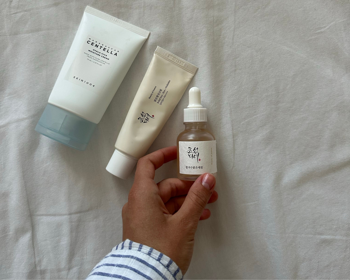 A hand holding a small dropper bottle of Beauty of Joseon serum, with a SKIN1004 Centella cream tube and a Beauty of Joseon sunscreen tube resting on a white fabric surface.