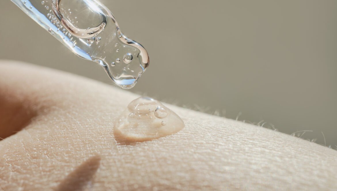 A close-up shot of a glass dropper dispensing a clear drop of serum onto a person's skin.