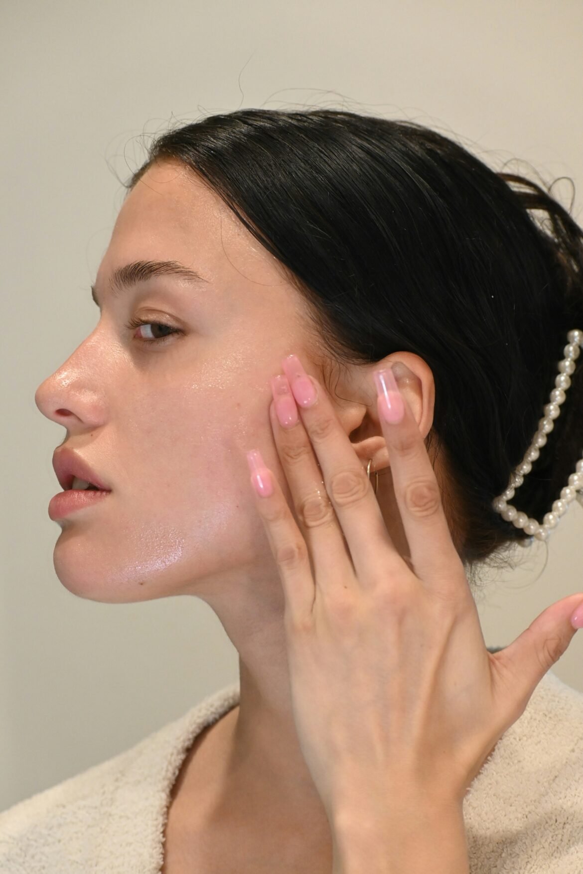 A profile view of a person with glowing, dewy skin wearing a white robe, gently pressing skincare products into their cheek to demonstrate a skincare routine.