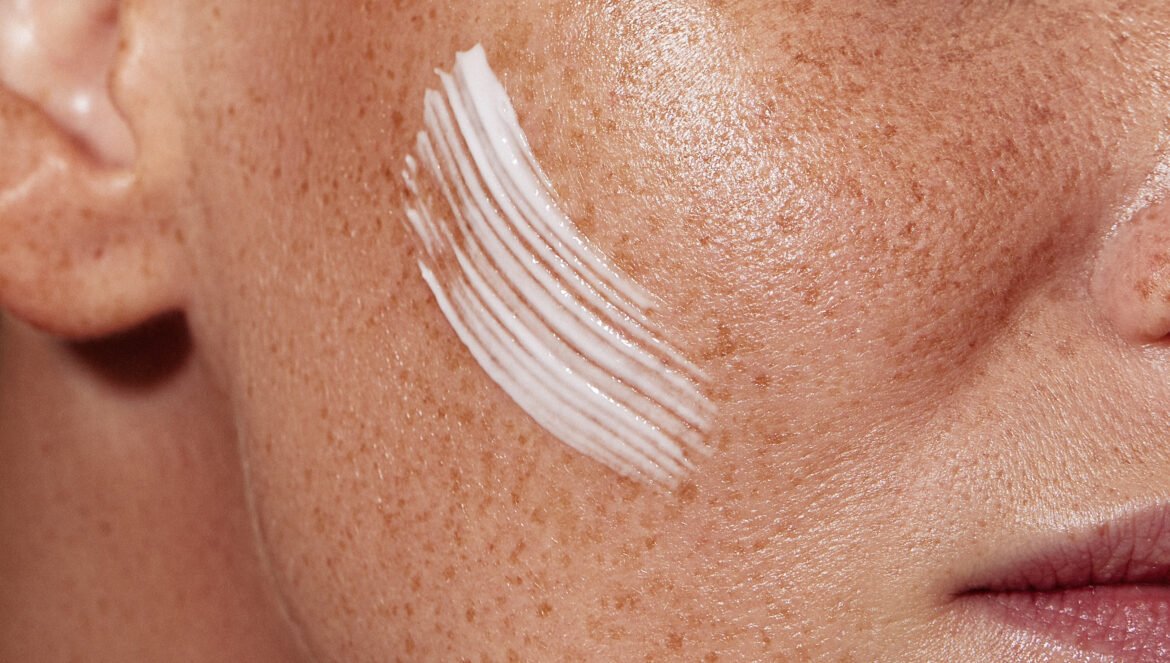 A close-up shot of a person's healthy, freckled skin with a thick, textured swipe of white skincare cream applied across their cheek.