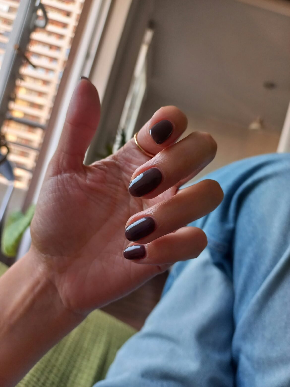 A close-up of a hand displaying a glossy, dark chocolate espresso manicure on short, rounded nails, paired with a thin gold ring and blue denim jeans.