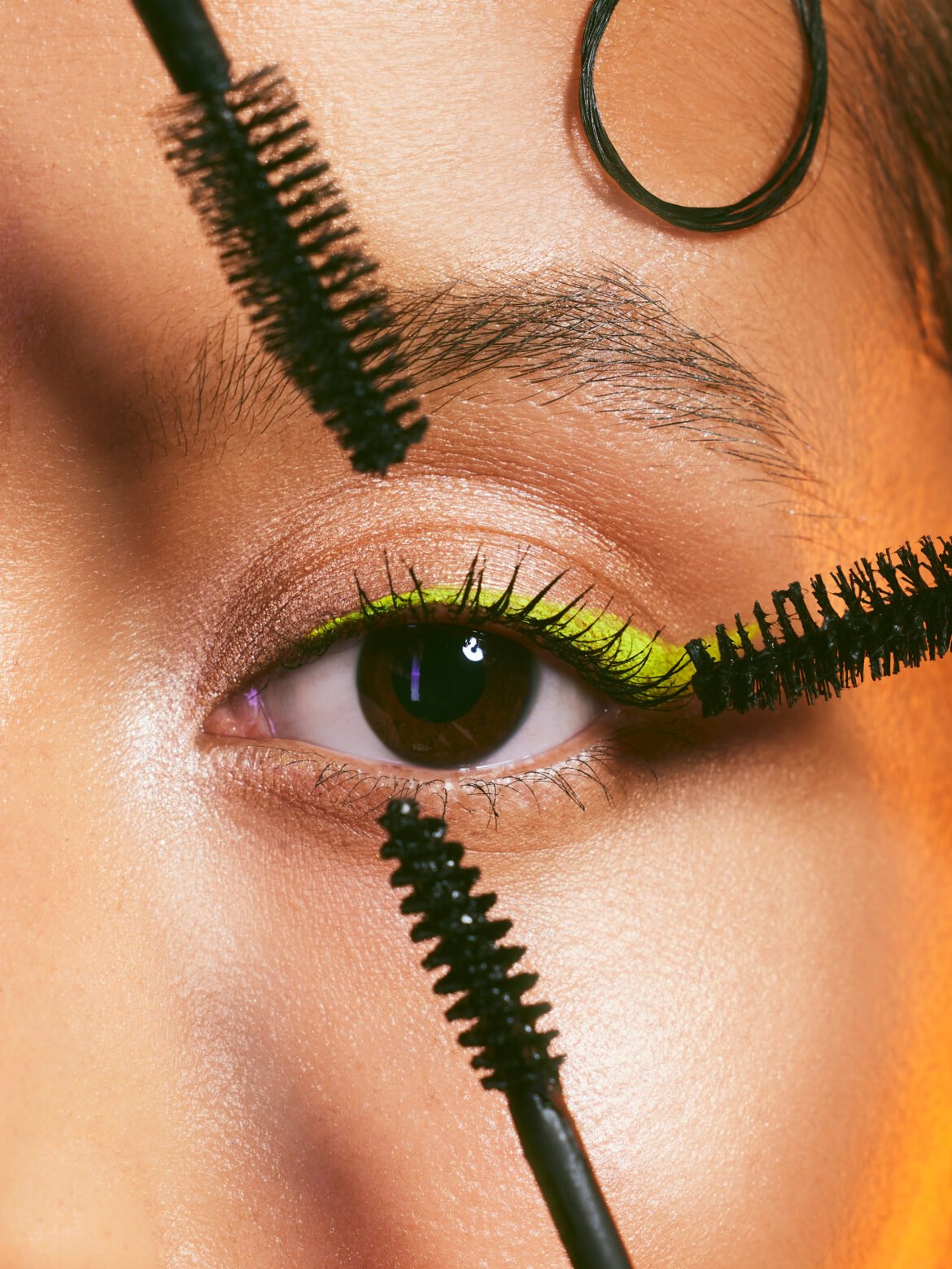 A close-up of a brown eye with neon yellow eyeliner and shimmery eyeshadow, as a wand applies black waterproof mascara. Text overlay reads "Best Waterproof Mascaras: 7 Top-Rated Formulas That Won't Budge."