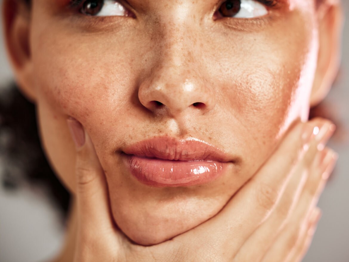 A close-up photograph focusing on the lower face of a person with natural, freckled skin. Their lips are coated in a high-shine, glossy pink product. Their hands are gently framing their face, touching their cheeks and chin.