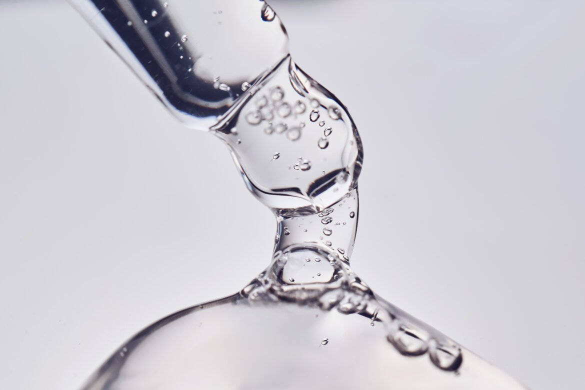 A macro close-up of a glass pipette dispensing a thick, clear cosmetic serum. The viscous gel stretches from the dropper tip to a pool below, with tiny air bubbles suspended inside the liquid.