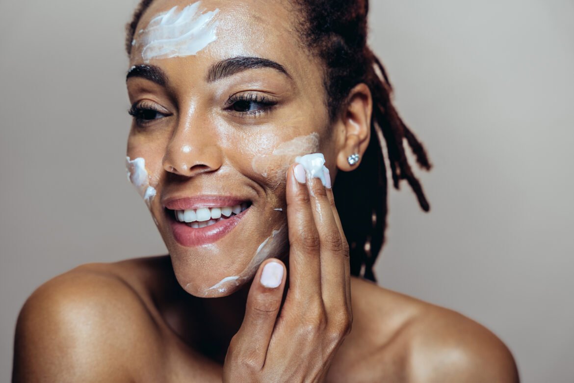 A smiling woman applying white face cream to her cheek in a home setting.