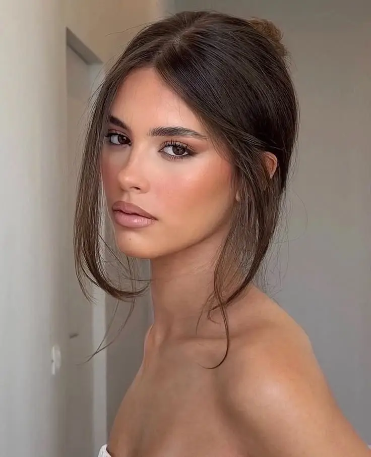 Close-up portrait of a woman wearing the toasty makeup trend with warm bronzed skin, earthy brown eyeshadow, and a neutral brown lip against a light background.