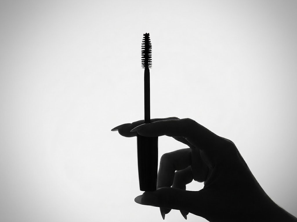 A black and white silhouette of a female hand with long, oval nails centered in the frame, delicately holding a mascara wand upright against a smooth, bright background.