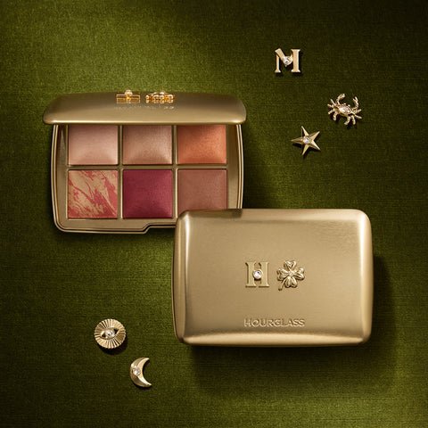 A custom gold Hourglass Ambient Lighting Edit face palette resting on dark olive green velvet. One palette is open displaying six baked powders, while a closed compact features an 'H' and clover charm, surrounded by scattered gold charms including a moon, star, and crab.