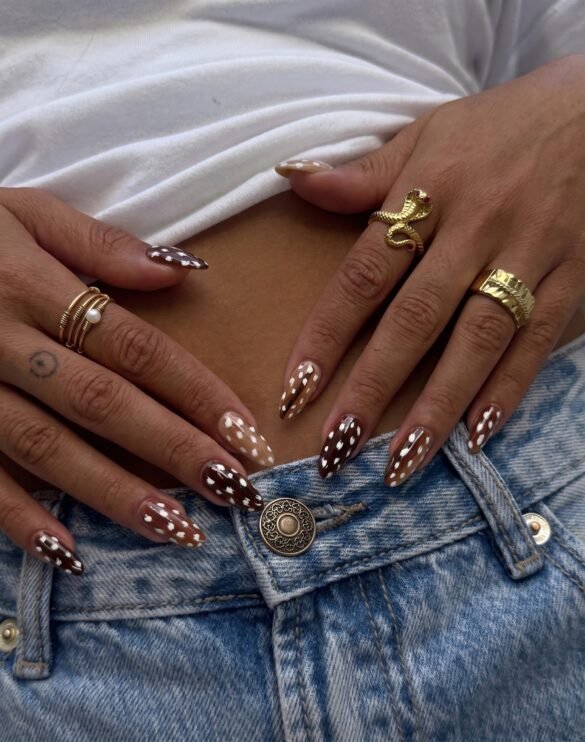A close-up photograph focuses on two hands with intricately detailed almond-shaped nails resting on the open waistband of blue denim jeans. The nails feature a rich dalmatian-patterned design in shades of brown, tan, and white. The hands wear several pieces of gold jewelry, including a coiled snake ring on one index finger and stacked rings with pearl accents on the other. A white t-shirt is visible at the top.