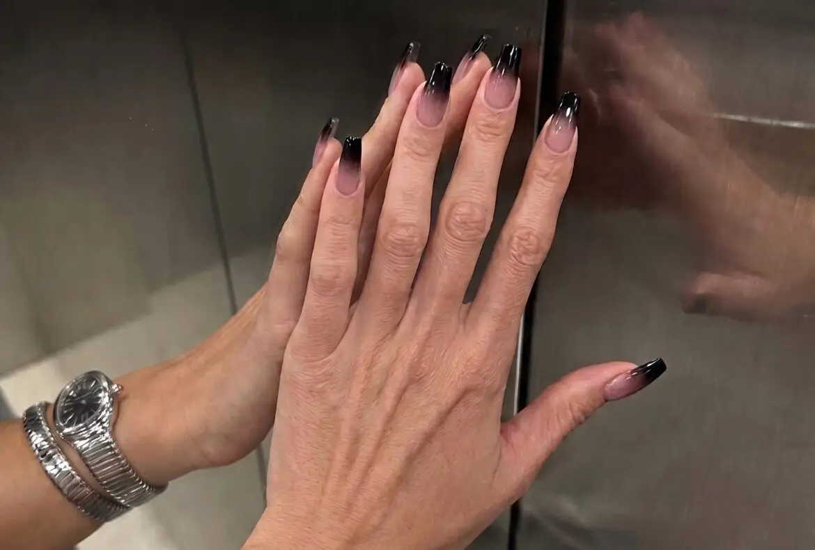 A detailed close-up photograph taken inside a brushed metal elevator. A person presses their right hand with long, square-tipped black ombre manicured nails against a mirrored surface. Their left hand is also visible, with fingers touching the reflections. Matching silver coiled snake-style watch bracelets are worn on both wrists. Reflections of the hands and jewelry are prominent.