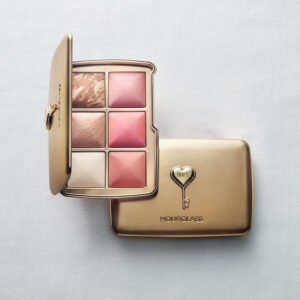 A gold Hourglass limited-edition Hope Palette from the 2026 Holiday collection. An open compact displaying six ambient lighting face powders rests on top of a closed compact featuring a raised heart-shaped key emblem.