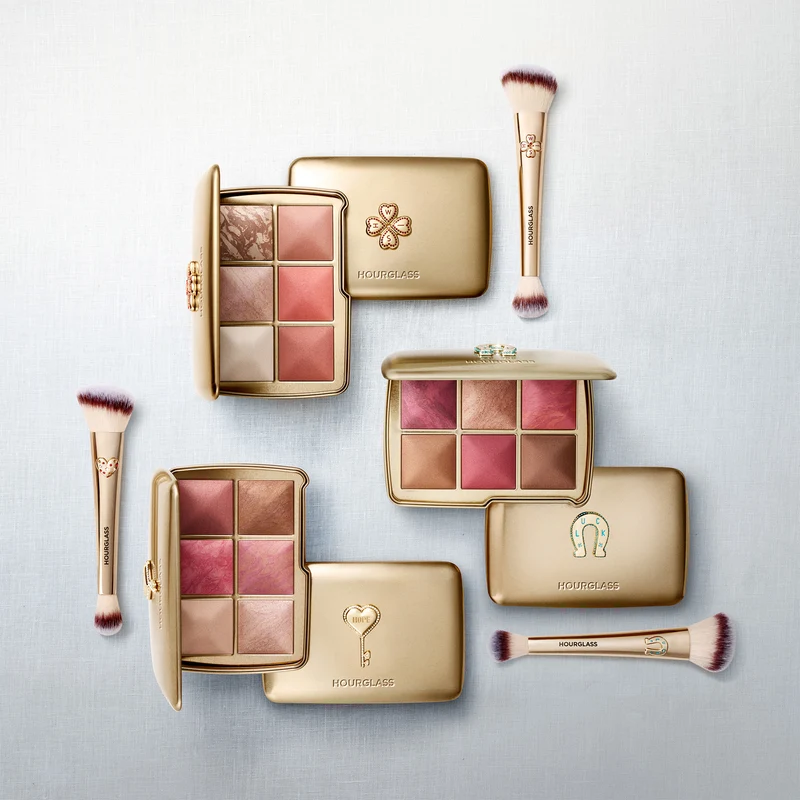 Three open Hourglass limited-edition 2026 Holiday Ambient Lighting Palettes in gold packaging, each displaying six face powders, arranged alongside three double-ended Hourglass makeup brushes on a light textured background.