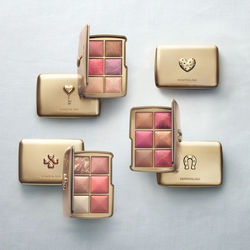 A flat lay of six gold Hourglass limited-edition holiday face palettes. Three are open, revealing six pans of luminous blush, bronzer, and highlighter, while three closed palettes display raised heart, key, and clover designs on a light background.