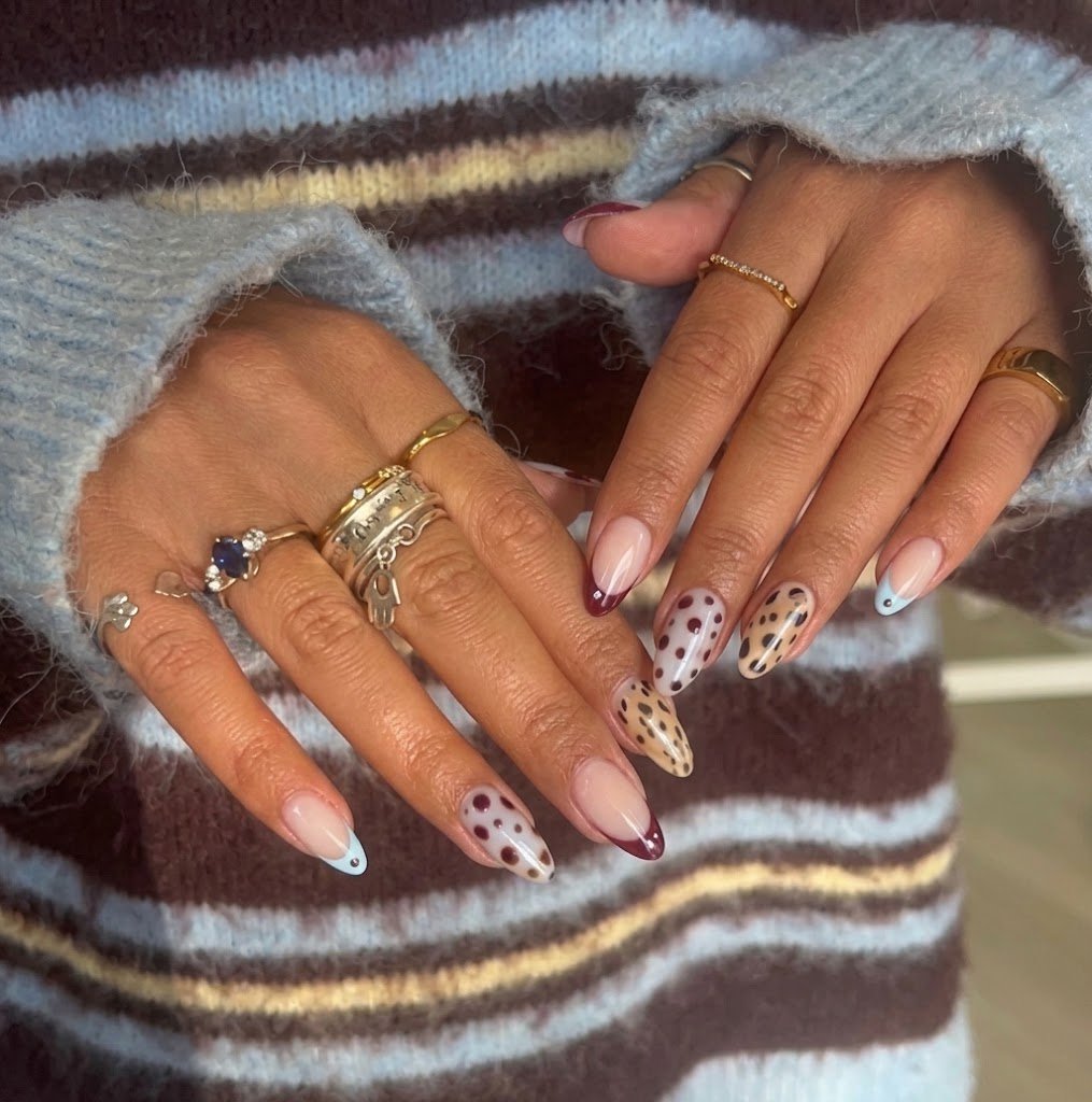 A detailed close-up view of two manicured hands resting against the cuff of a striped brown and light blue knit sweater. The almond-shaped nails display an eclectic, mix-and-match fall manicure featuring alternating designs of blue micro French tips, deep maroon polka dots on a milky base, and natural leopard print spots. The fingers are adorned with multiple stacked gold and silver rings, including a hamsa ring and a sapphire ring.