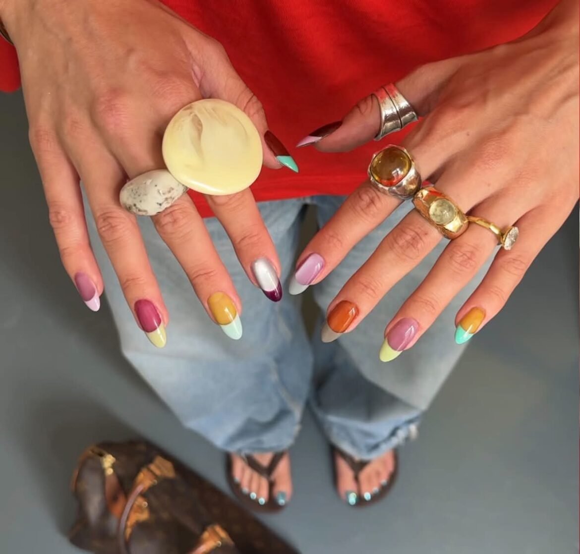 A top-down view of hands showcasing a colorful mismatched jelly French manicure on almond-shaped nails, accessorized with chunky statement rings.