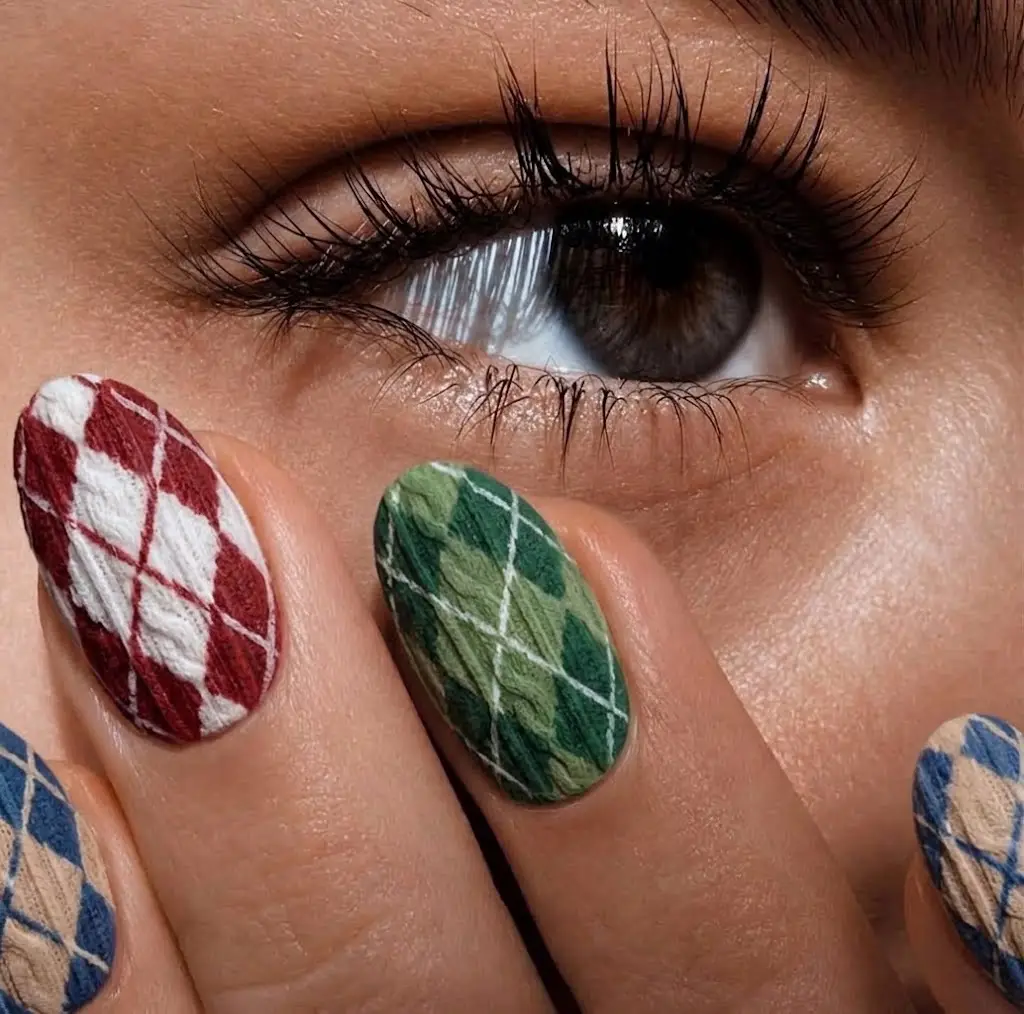 A close-up of a brown eye with long, dark eyelashes. In the foreground, fingers are resting against the skin under the eye. The nails are decorated with a textured, three-dimensional argyle pattern (diamond pattern) that looks like knitted yarn. The visible nails have different color combinations, including red and white, green and light green, as well as blue and beige.