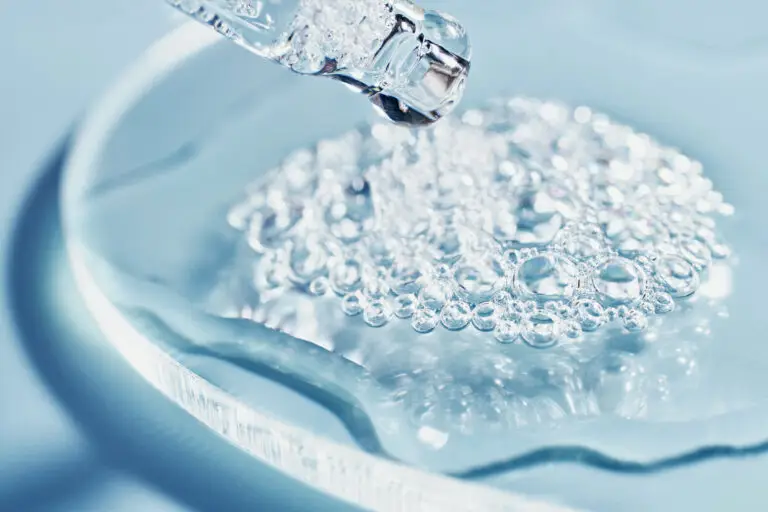 A close-up photograph of a clear pipette dispensing a clear liquid, filled with small air bubbles, onto a glass watch glass. The scene is on a light blue background with a natural shadow.