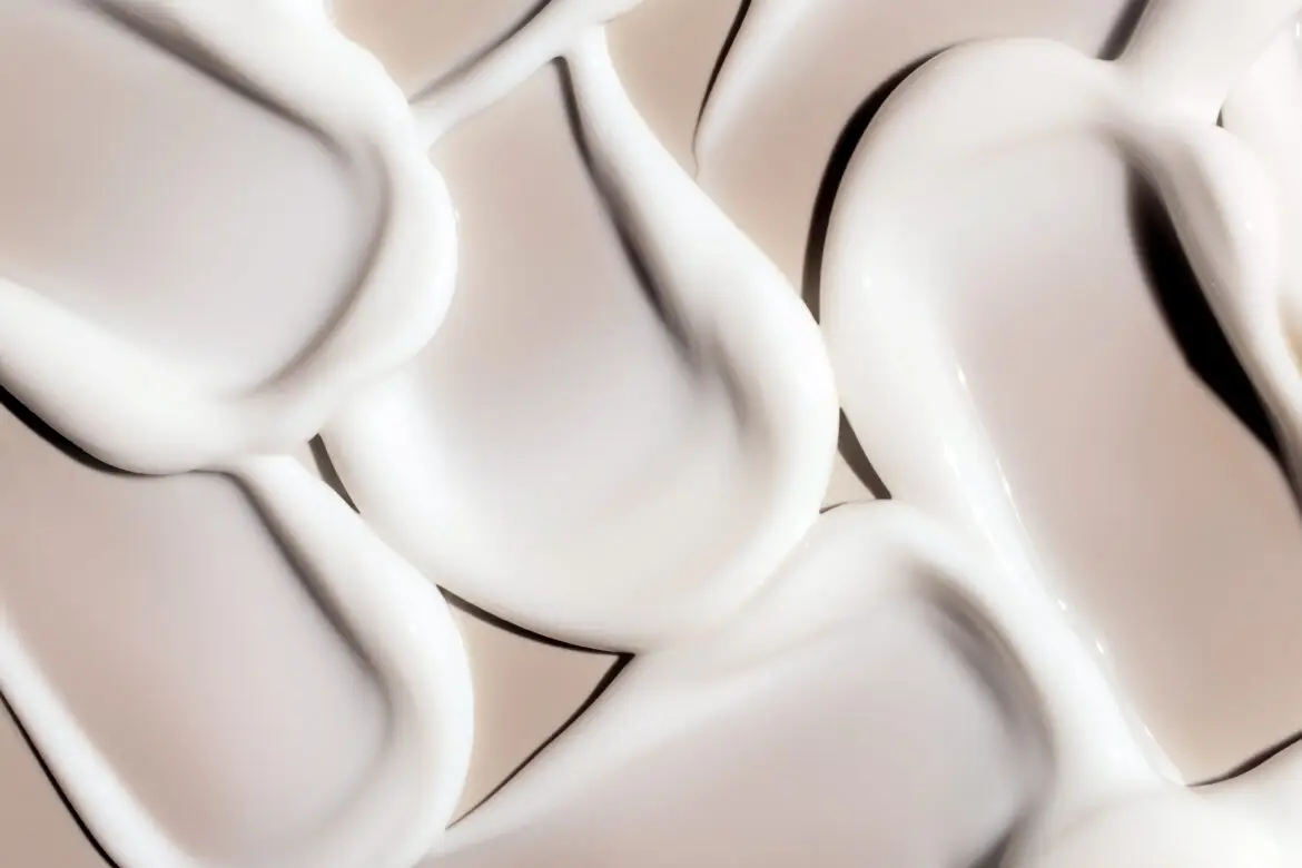 An abstract macro photograph of multiple thick, sculpted swatches of smooth, white cosmetic cream with defined edges and a glossy finish, overlapping against a warm taupe background.