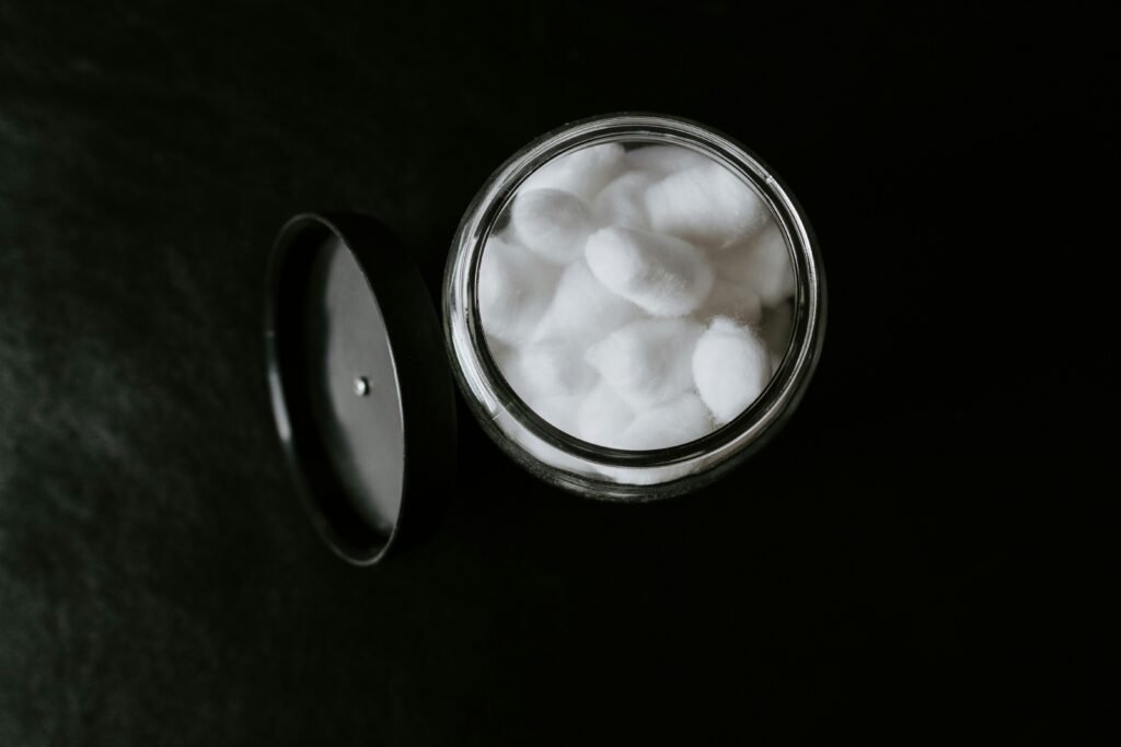 Top-down view of soft white cotton balls stored in a clear glass jar on a dark background, essential for applying facial toner and daily makeup removal.