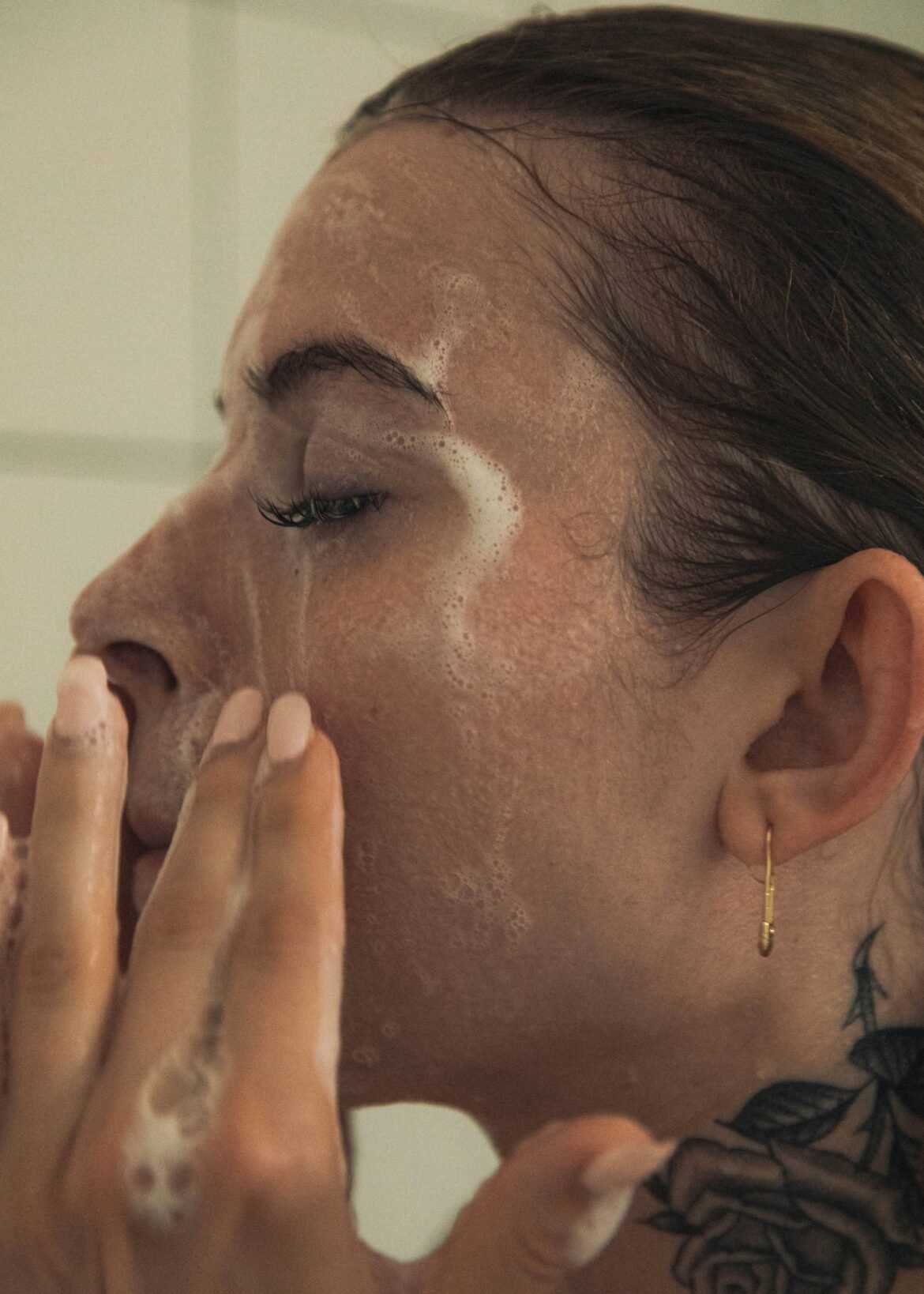 Close-up profile of a person washing their face with a foaming cleanser. They are gently massaging the lather into their wet skin with their fingers, with their eyes closed.