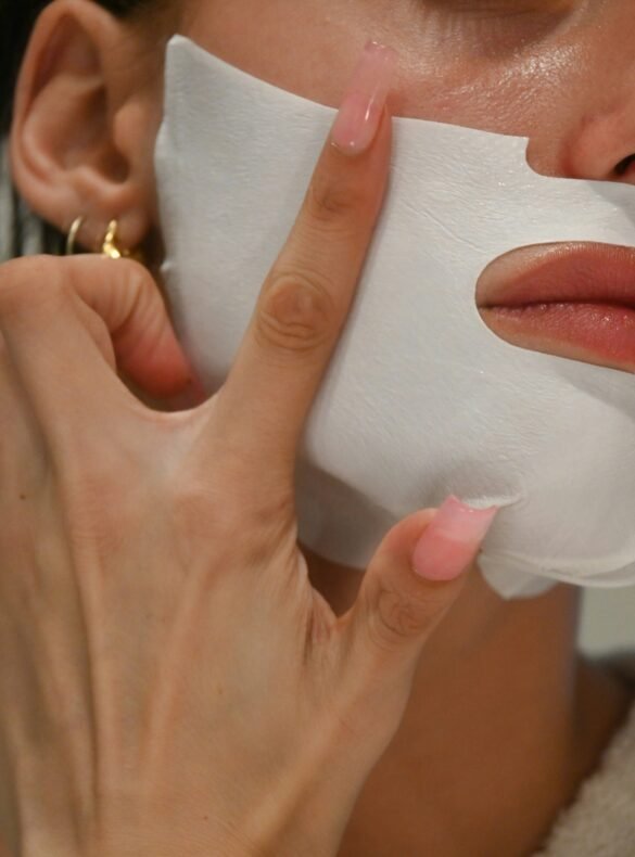 A close-up of a person's hand with long, light pink square nails gently pressing a white sheet mask onto their cheek. The person has glossy lips and is wearing small gold hoop earrings.