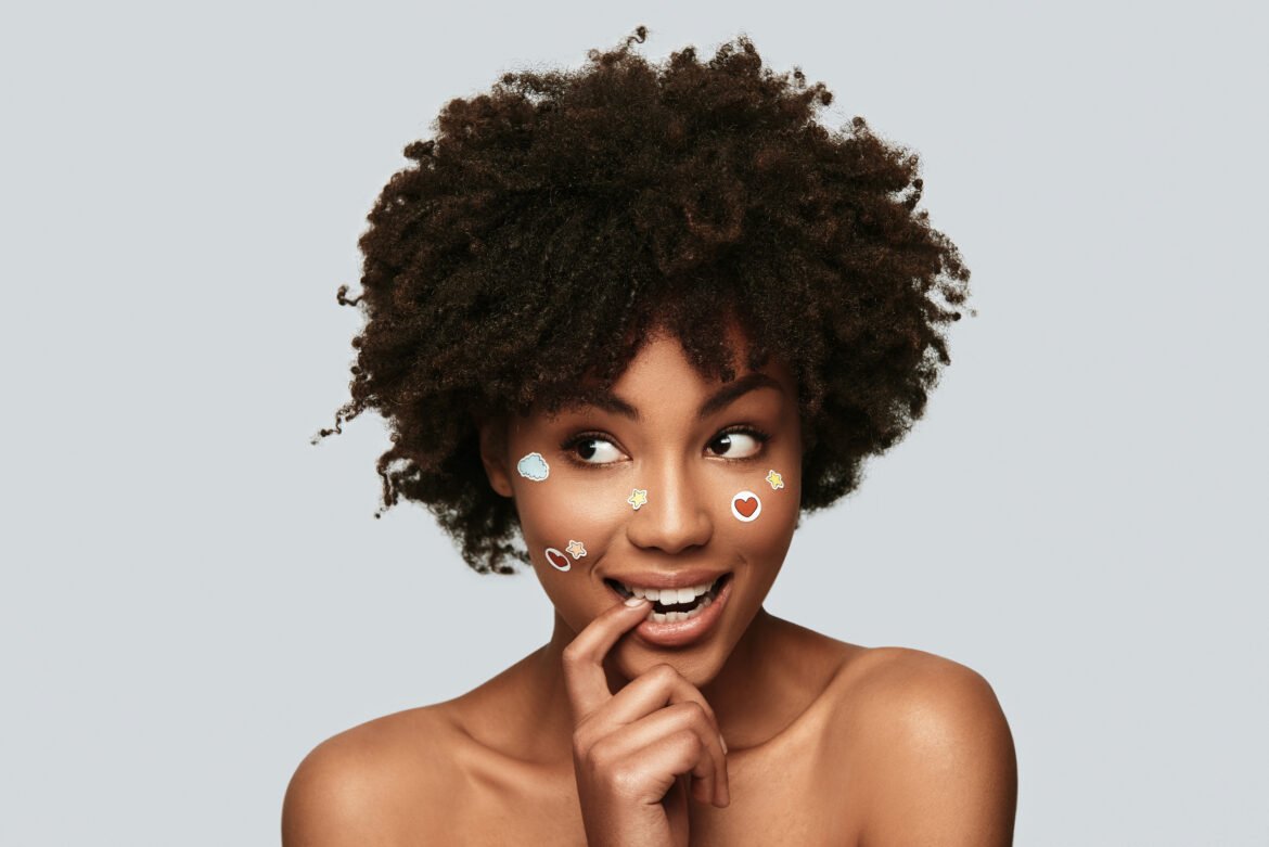 A studio portrait of a Black woman with a natural afro and bare shoulders against a light gray background. She has a playful expression, looking to the side with her finger at her mouth, and is wearing small, colorful cartoon stickers (like a cloud and a heart) on her cheeks.