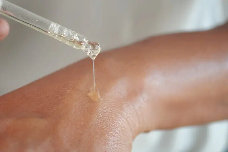 A close-up photograph of a clear, viscoelastic serum being precisely applied to the textured back of a mature hand using a glass pipette dropper. A single drop is caught just as it makes contact with the skin, with a fine string of the product visible.