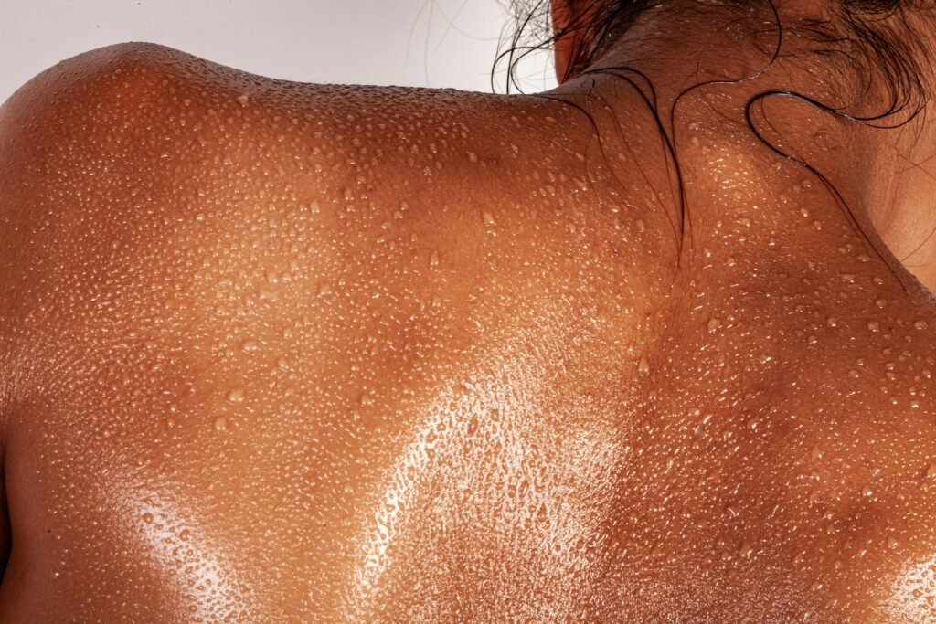 Close-up of glowing, bronzed skin on a person's bare back and shoulders, covered in glistening water droplets with wet hair at the nape of the neck.