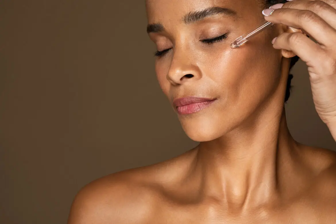 A Black woman with glowing skin and closed eyes applying a clear serum to her cheek using a glass dropper against a warm brown background