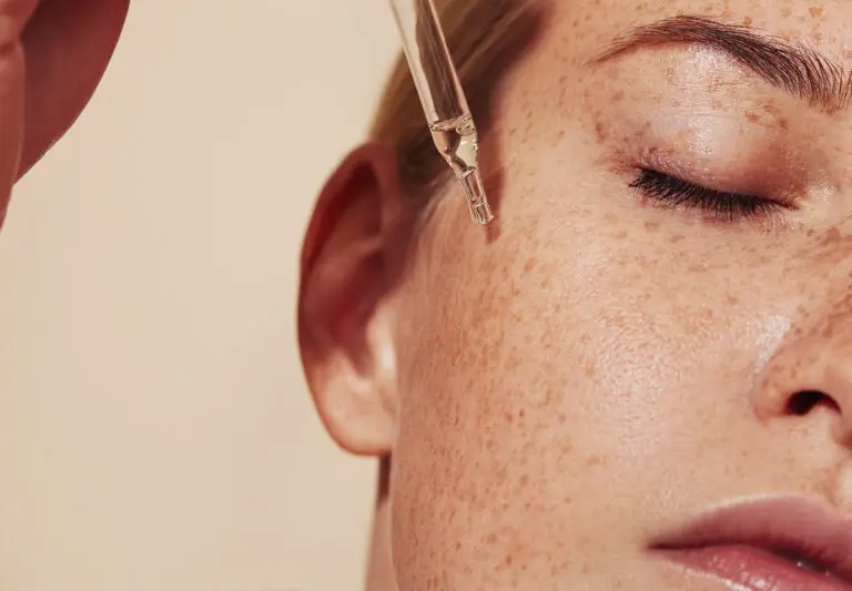 A close-up photograph of a young woman with natural freckles across her cheek and around her closed eyes. A hand is holding a glass dropper above her face, with a single clear serum droplet about to fall toward her cheek.