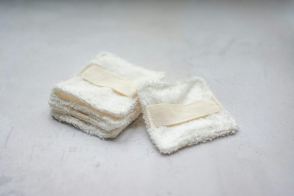 A stack of reusable white terry cloth makeup remover pads with beige finger straps, resting on a light grey textured background.
