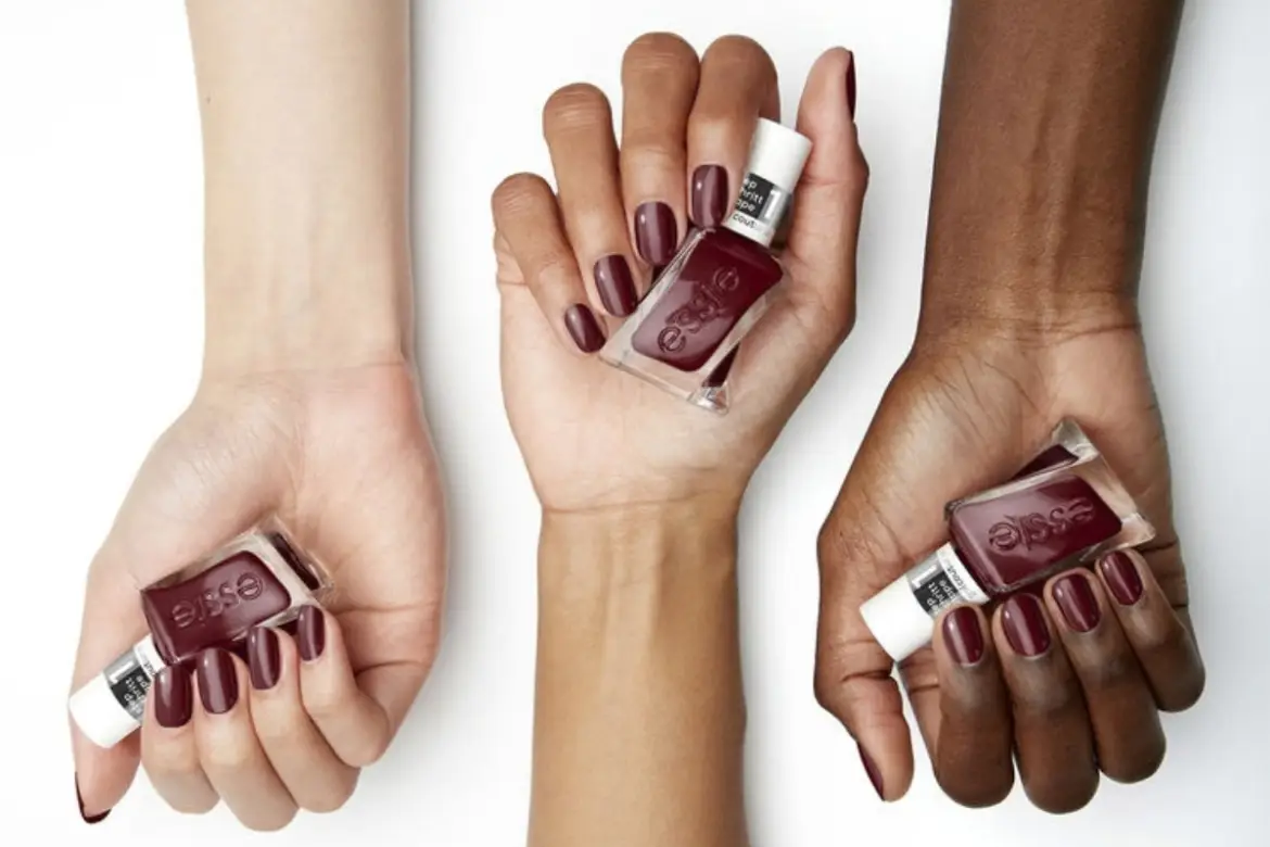 Three hands with varying skin tones holding bottles of Essie burgundy nail polish against a clean white background, showcasing the cherry noir manicure trend.